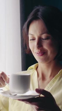 Kadın bir kahve fincanı tutuyor ve sabah ışığında pencereden dışarı bakarken gülümsüyor..