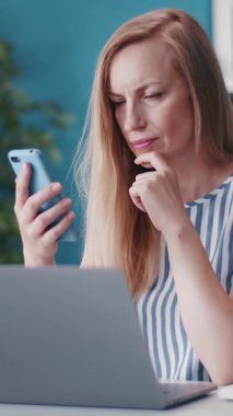 Kadın akıllı telefonu kontrol ederken yanında dizüstü bilgisayarı olan bir masada oturuyor..