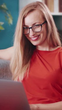 Bir kadın gün içinde çalışırken ve gülümserken dizüstü bilgisayar kullanarak kanepede oturur..