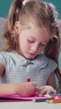 Bir kız kırmızı ve pembe kalemlerle kapalı bir alanda kağıt üzerine resim çizer..