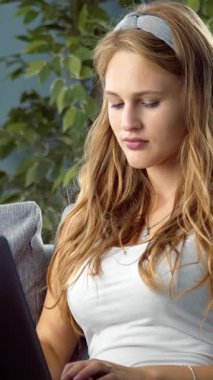 Kadın dizüstü bilgisayarla kanepeye oturuyor, ev ortamında ekrana odaklanıyor..