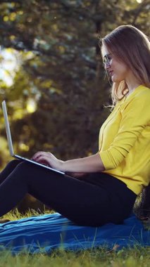 Sarı bluzlu bir kadın parkta mavi bir battaniyede dizüstü bilgisayar kullanıyor..