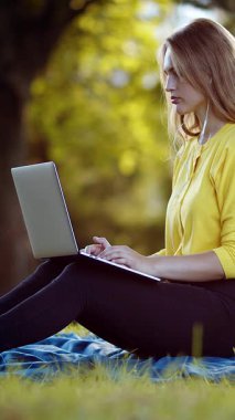 Kadın, parktaki ağaçların altında dizüstü bilgisayar kullanarak battaniyeye oturur..