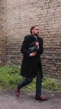 Siyah paltolu bir adam elinde bir çanta ile tuğla bir duvara doğru koşuyor..