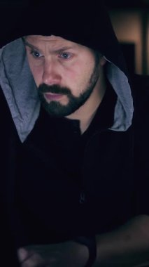 Karanlık başlıklı bir adam yoğun bir şekilde düşük ışıkta dizüstü bilgisayarda çalışır..