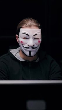 Maskeli bir adam karanlık bir odada bir masada oturur ve gece bilgisayar kullanır..