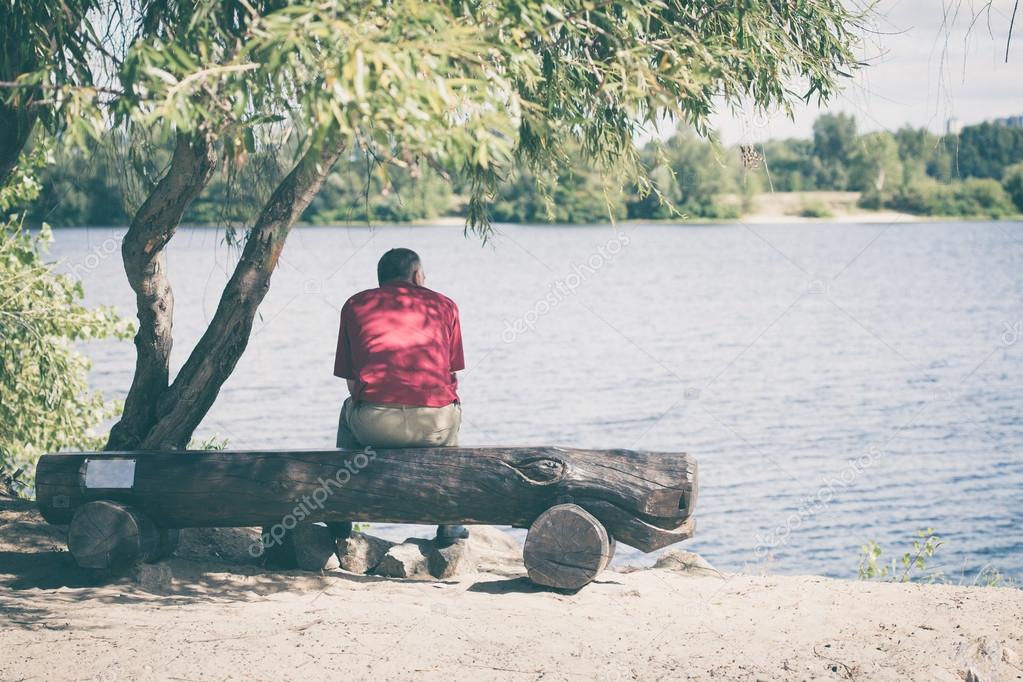 Old man sitting on a bench by the river - Stock Photo, Image. 