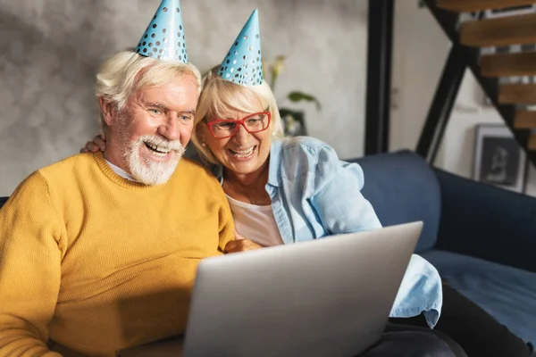 Cheerful modern caucasian elderly couple making video call on laptop for birthday or anniversary celebration while sitting on sofa at home. Videoconference holiday greetings concept