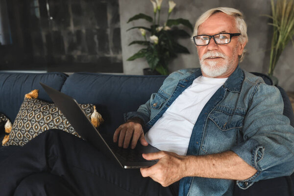 Elderly man with glasses with gray hair and a beard sits at home on the sofa in and works or browses social networks on a laptop looking at the camera