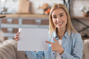 Kafkasyalı mutlu kadın vlogger gülümsüyor ve elinde boş bir kağıt tutarak kanepede oturmuş video kaydediyor.