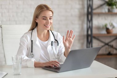 İş yerindeki beyaz önlüklü genç bayan doktor, video çağrısına göre hastayı selamlıyor