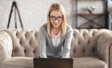 Evdeki kanepede oturan güzel sarışın kadın mutlu yüzlü bilgisayar laptopu kullanarak çalışıyor.