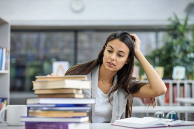 Güzel kız öğrenci üniversite kütüphanesinde masada otururken kitap yığınına bakar ve başını tutar. Öğrenim ve eğitim kavramı