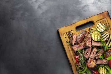 Dilimlenmiş orta boy biftek, ızgara sebzeler, ahşap kesme tahtası, koyu arka plan, fotokopi odası manzaralı.