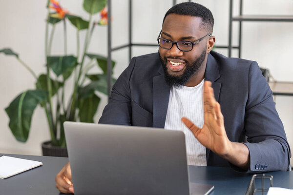 Happy African American businessman at video conference, talking with colleagues about work process using laptop, sitting at the desk, smiling and waving hand