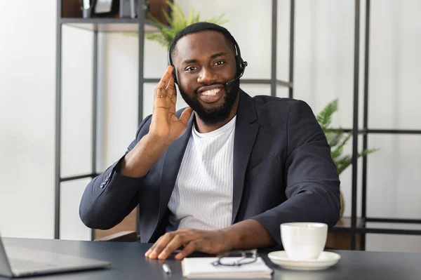 Happy African American man in headset and suit looks at the camera, smiling. Successful male operator of call center sits at the table in office. Consulting people concept