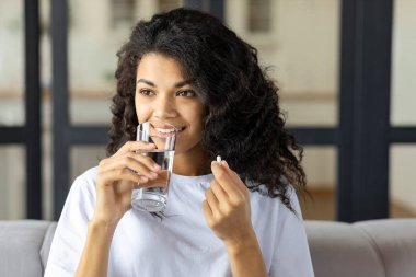 Elinde vitamin hapı ve bir bardak su tutan güzel, gülümseyen Afro-Amerikan kadın evdeki kanepede oturuyor. Sağlıklı yaşam tarzı, sağlıklı beslenme konsepti