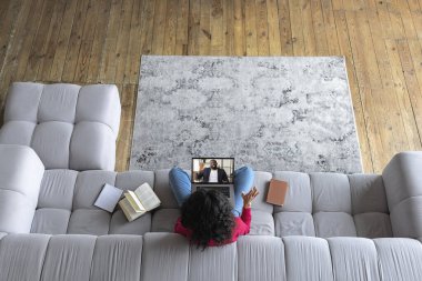 Top view 'de genç bir kadın evinde bir öğretmen ya da arkadaşıyla kanepede otururken çevrimiçi konferans ya da video görüşmesi yapıyor. Çevrimiçi eğitim veya uzaktan çalışma kavramı