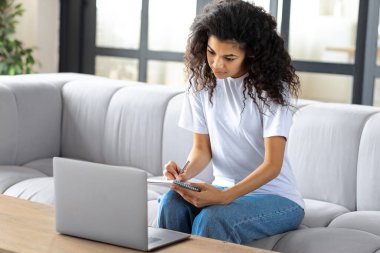 Genç Afrikalı Amerikalı kız notlar alıyor, çalışıyor, online ders alıyor ya da web sitelerine giriyor. Ev kavramında çevrimiçi öğrenim