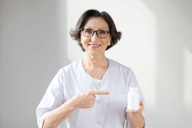 Beyaz önlüklü gülümseyen olgun kadın doktor portresi beyaz arka planda ilaç şişesi taşıyor.