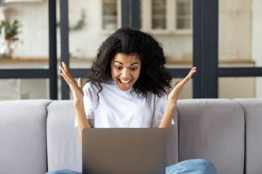 Evdeki koltukta oturan mutlu Afrikalı Amerikalı kadın mutlu bir şekilde piyangoyu kazandı ya da laptopta harika haberler aldı.