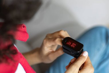 Afro-Amerikalı kadın nabız ve oksijen doygunluğunu evdeki kanepede oturan nabız oksimetresiyle ölçüyor. Sağlık sigortası kavramı.