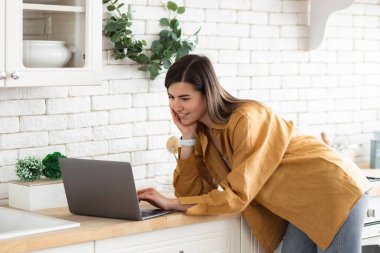 Genç bayan dizüstü bilgisayar kullanıyor ve evde mutfakta çalışıyor. Sosyal ağlarda bilgisayar kullanarak iletişim kuran güzel bir kadın.