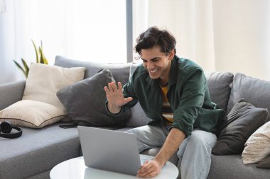 Mutlu başarılı, genç, Kafkasyalı, ofisteki kanepede dizüstü bilgisayarla oturmuş video konferansında günlük kıyafetlerle konuşuyor.