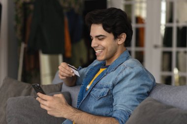 Elinde plastik kredi kartı ve akıllı telefon tutan gülümseyen beyaz adam uzaktan para ödediği için internetten alışveriş yapıyor ve banka hizmetleri üzerinden güvenli ödeme yapıyor.