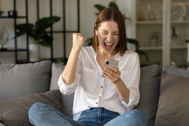 Mutlu beyaz kadın cep telefonuyla iyi haber alıyor, başarıyı kutluyor, evdeki koltukta oturuyor.