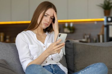 Kanepede oturan hayal kırıklığına uğramış genç beyaz kadın akıllı telefonda beklenmedik kötü haberler karşısında hayal kırıklığına uğradı.