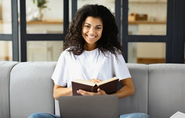 一个年轻的非洲裔美国女人坐在沙发上 拿着一本书和笔记本电脑 看着相机 开心地微笑着 网上教育的画像 — 图库照片