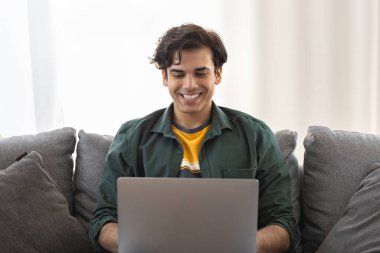 Genç, neşeli bir adam dizüstü bilgisayarla koltukta oturmuş bilgisayar monitörüne bakıyor ve gülümsüyor. Yakışıklı genç adam sosyal medyada komik videolar izlemekten hoşlanıyor