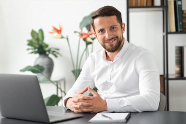 Ofisteki çalışma masasında dizüstü bilgisayarla oturan başarılı genç iş adamının portresi. Beyaz erkek işçi iş yerinde otururken kameraya bakıyor.