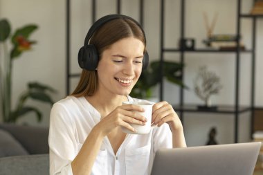 Kulaklıkla kahve içip gülümseyen beyaz kadın çalışanlar iş arkadaşlarıyla görüntülü konuşma yapıyor, genç bir kadın bilgisayarında internet konferansı yapıyor.