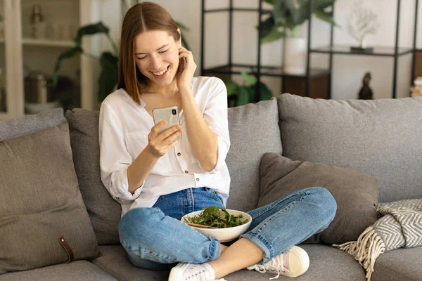 Smiling young caucasian millennial woman chatting in social networks using mobile applications sitting on couch at home