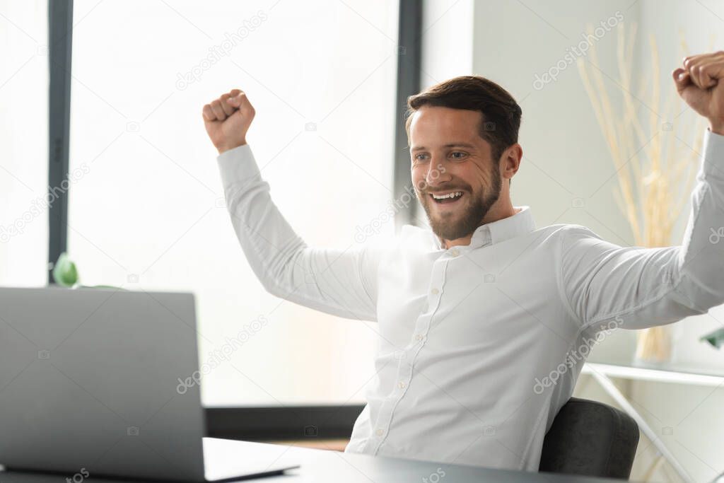 Excited young caucasian man celebrating dream goal achievement. Successful businessmen rejoices financial project results sitting at the desk in the office