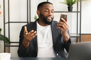 Başarılı bir Afrikalı Amerikalı iş adamı cep telefonuyla konferans yapıyor. Ofiste oturan meslektaşlarıyla, müdürle, akıllı fondaki ortak çalışma alanı konuşmalarında oturuyor.