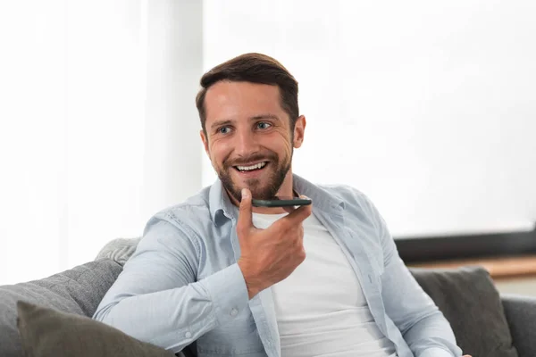 Caucasian bearded guy holding a mobile phone in his hand, talking on a speakerphone or audio message recording on a mobile phone sitting at home on the couch, looking away, smiling