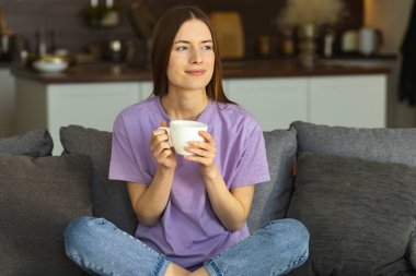Kafkasyalı mutlu bir kadın elinde bir fincan kahveyle evdeki kanepede dinleniyor. Hafta sonunun tadını çıkaran genç bir kadın, başka tarafa bakıyor ve geleceği düşünüyor, tatlı tatlı gülümsüyor.