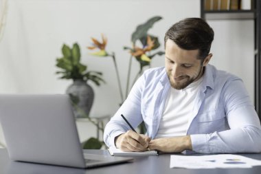 Yoğunlaşmış beyaz adam evde veya ofiste dizüstü bilgisayarla masaya otururken not alıyor, genç işadamı yeni işi için bir kalkınma planı yazıyor, internetten öğrenim görüyor, e-öğrenim görüyor.
