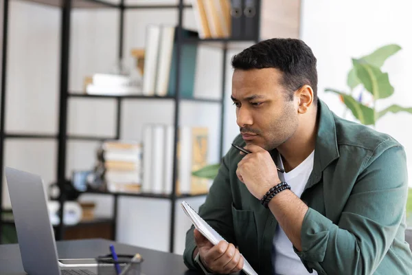 Concentrated Indian man looking at laptop screen while sitting at home office, analyze work, watch business webinar, online education