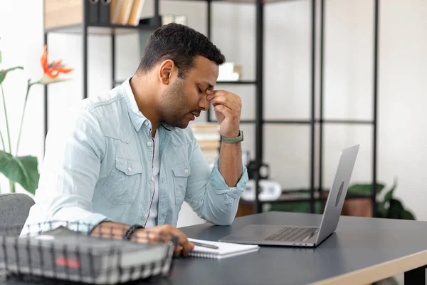 疲倦的印度商人坐在写字台前头疼或偏头痛 年轻的自由职业者坐在家里的写字台前 工作负担过重 眼睛因电脑而感到疲劳 — 图库照片