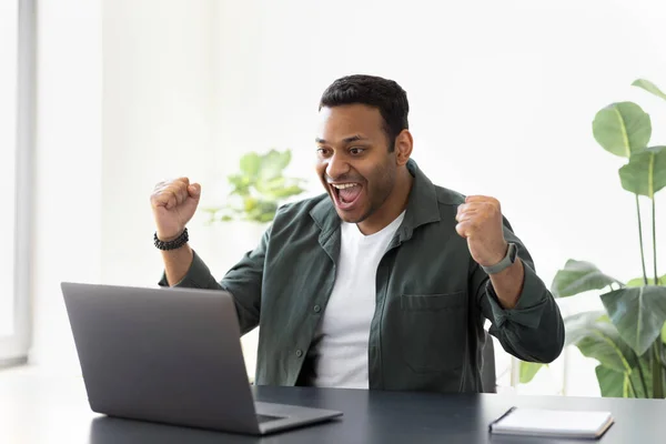 Excited young Indian man celebrating dream goal achievement sitting at a desk in a home office or received good news on email