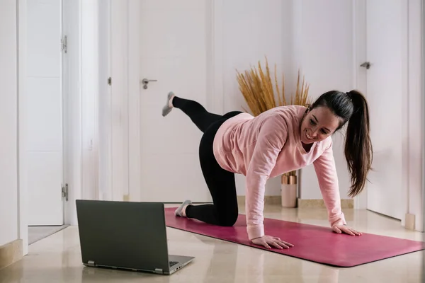 Koyu renk saçlı beyaz kadın laptopta spor dersini izlerken evde antrenman yapıyor.