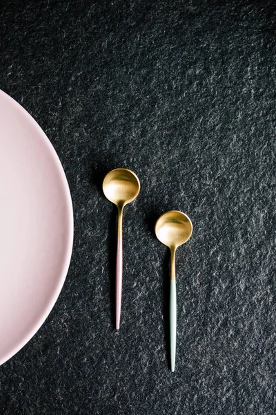 Siyah arka planda pembe bir tabağın yanında iki şık ve renkli kaşık. Boşluğu Kopyala