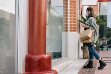 Sarı saçlı, koruyucu maskeli beyaz bir kadın alışveriş merkezinin vitrinine bakıyor. Bir elinde bir sürü alışveriş çantası taşır. Seçici odak