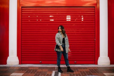 Beyaz kadın, sarı saçlı, kırmızı arka planda poz veriyor. Portre tam boy, yana bakıyor. Günlük kıyafetler ve siyah bir çanta giyiyor..