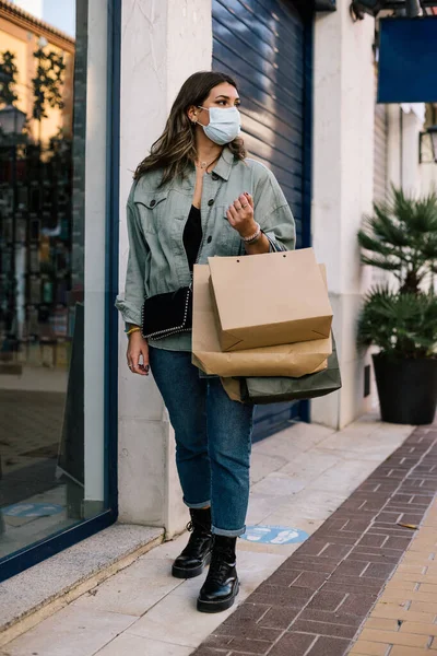 Yan tarafa bakarken alışveriş merkezinde yürüyen bir kadın. Alışveriş torbalarını elinde tutuyor. Sıradan bir elbise, oje, çanta ve maske takıyor.