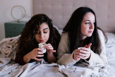 İki beyaz kadın bir bardak kahve tutarak yatakta uzanıyorlar. Biri pencereden dışarı bakar, diğeri de bardağını üfler.